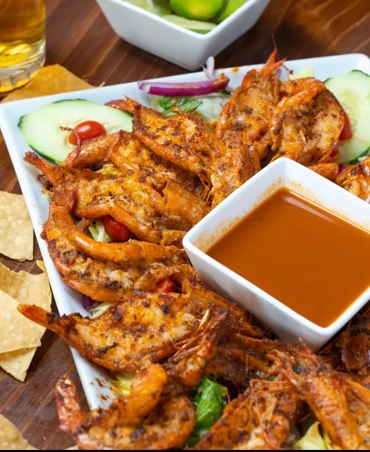 Un platón de camarones fritos con cáscara, con salsa anaranjada para acompañar, pepino y tomate.